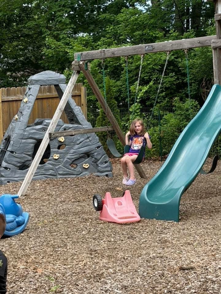 Child on a climbing wall and slide surrounded by green trees at Mother Goose Day Care
