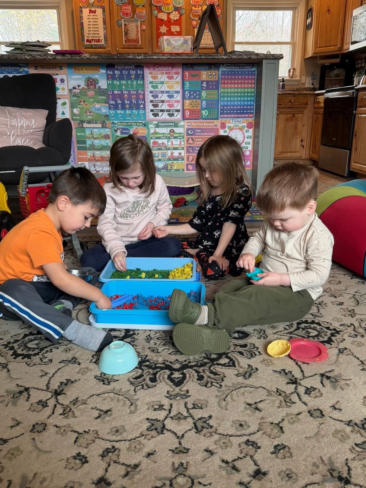 Four young children sitting on a patterned rug playing with a blue sensory bin at Mother Goose Day Care